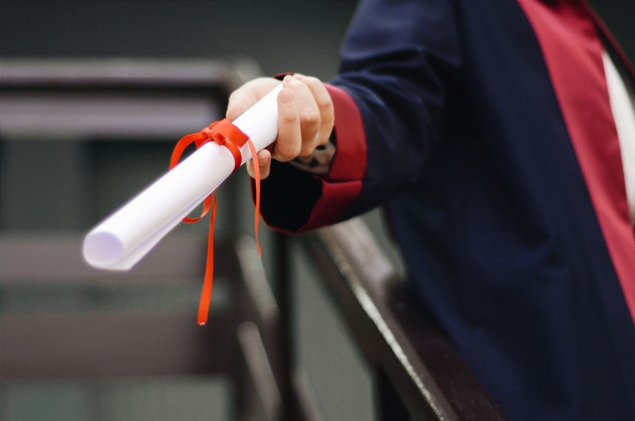 Home A close-up image of a graduate holding a diploma tied with a red ribbon, symbolizing achievement and success.