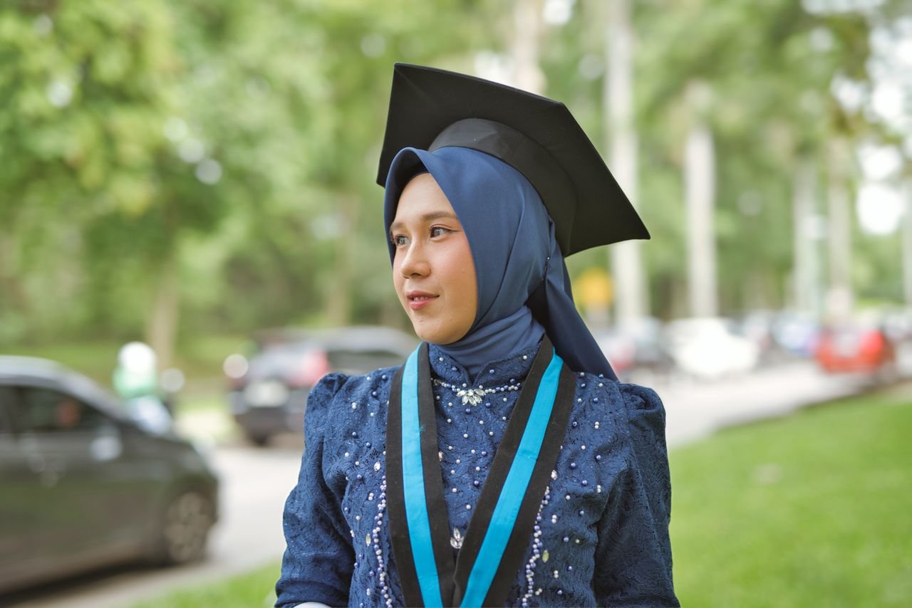 About Young woman in hijab wearing a graduation cap, celebrating achievement outdoors.