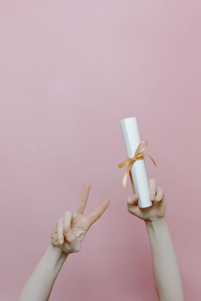 Home Diploma held in hand with peace sign on pastel pink background.