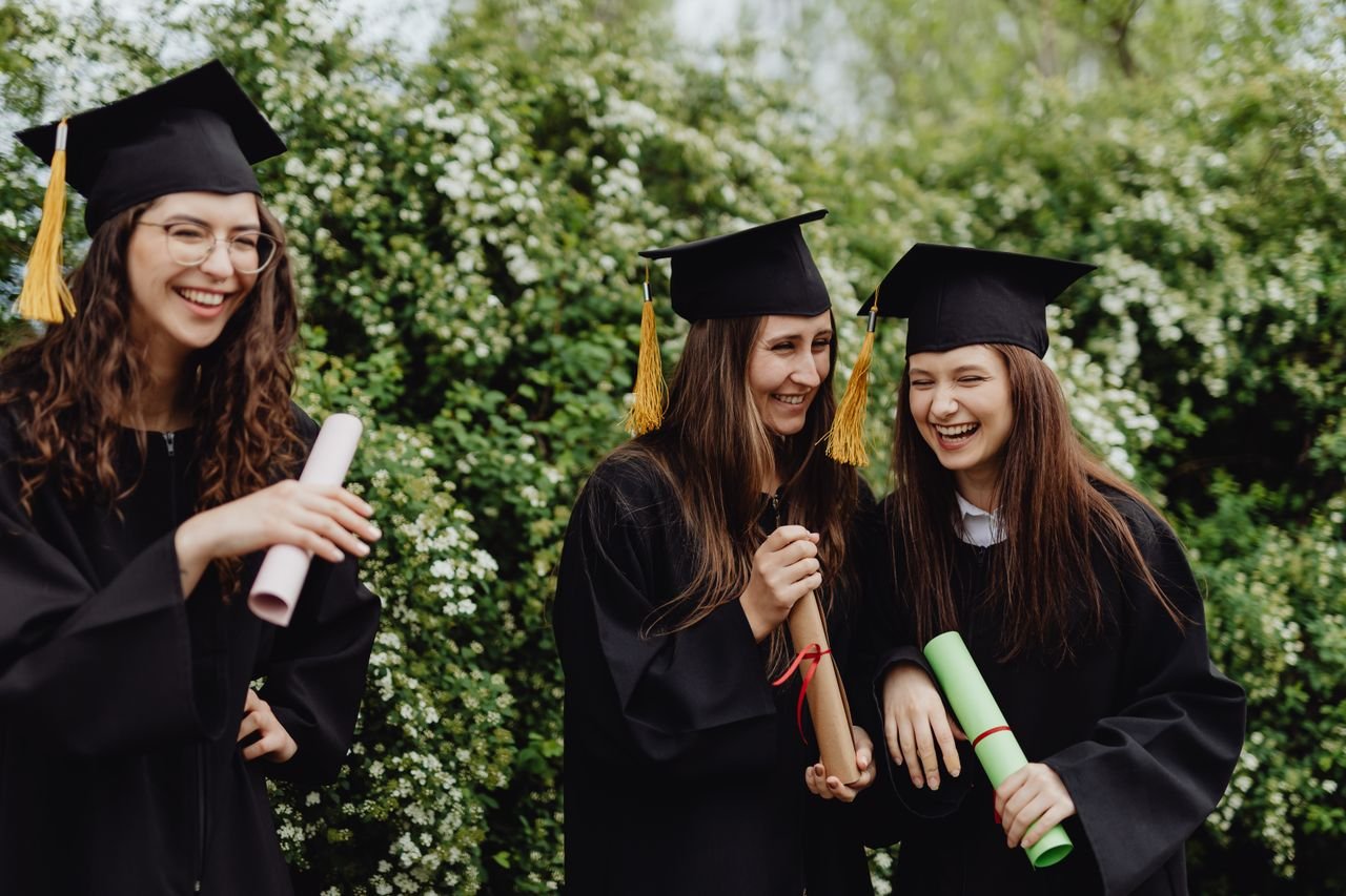 Home Group of happy female graduates in caps and gowns laughing and holding diplomas outdoors.