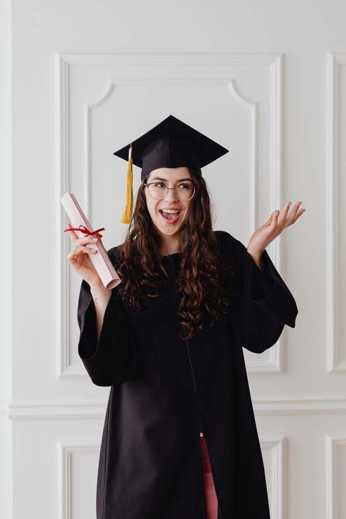 About Joyful young woman in graduation gown holding diploma, celebrating her achievement indoors.