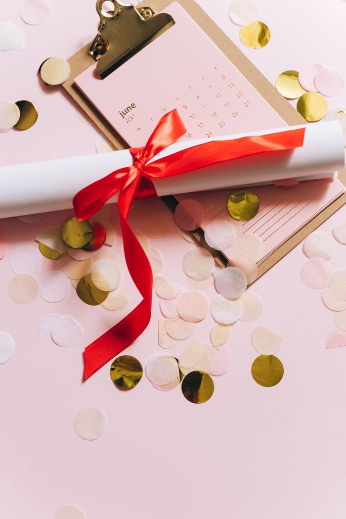 Study Abroad Flat lay of a rolled diploma with red ribbon on pink background with calendar and confetti.
